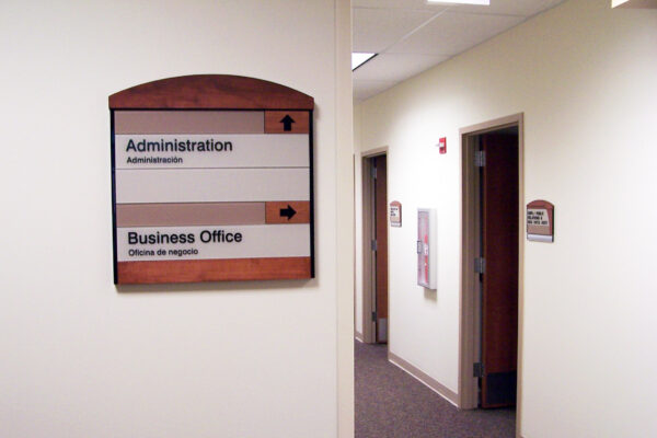 A hallway in an office building with beige walls and carpet. A sign on the left wall has two sections, one pointing up for “Administration” and one pointing right for “Business Office.” The hallway has several doors and is lit by ceiling lights.