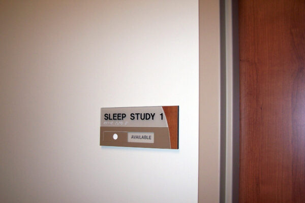 A sign on a light-colored wall reads "Sleep Study 1." Below the text, there is a small rectangular light with the word "AVAILABLE" illuminated in green. To the right of the sign is a wooden door.