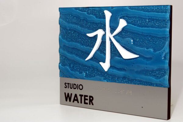 A sign with a blue background featuring textured, wave-like patterns and a prominent white Chinese character for "water." Below the blue section, there's a gray section with the words "STUDIO WATER" in black lettering.