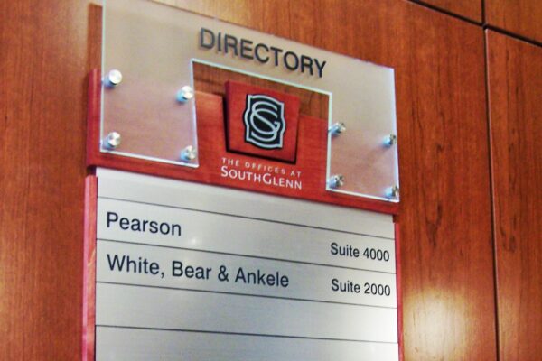 A wall-mounted directory sign with a clear header displaying the "University of South Glenn" logo. Below, it lists three entities: "Pearson" in Suite 4000, "White, Bear & Ankele" in Suite 2000, and "Z1 Solutions" in Suite 2500. The background is wooden.