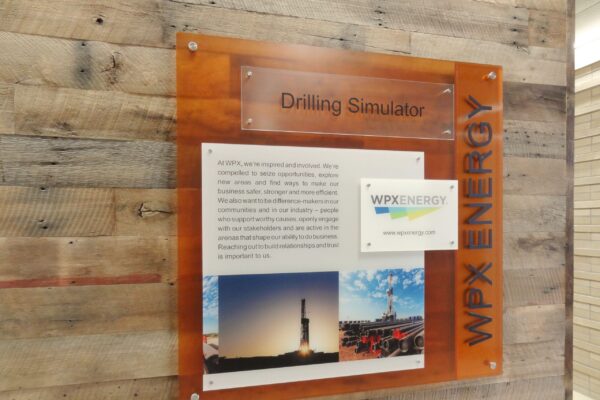 A sign mounted on a wooden wall reads "Drilling Simulator" at the top. Below, a description with the WPX ENERGY logo is displayed, along with two images: one of a drilling rig at sunset and another of a drilling operation. The WPX ENERGY logo is also on the side.