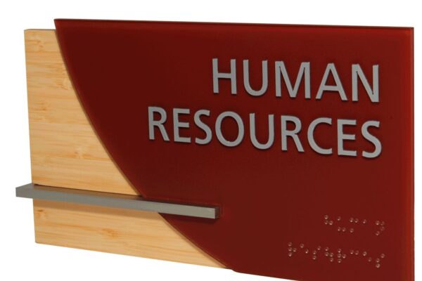 A wall-mounted sign with "Human Resources" written in large white letters on a red background. The sign also includes Braille text at the bottom. The sign has a wooden part on the left side with a horizontal metal bar extending halfway across.