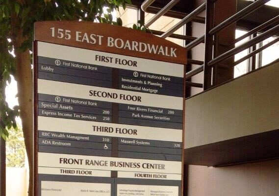 Directory sign at 155 East Boardwalk showing floor listings for various businesses, standing in an indoor setting with surrounding potted plants and a background of windows and railings.