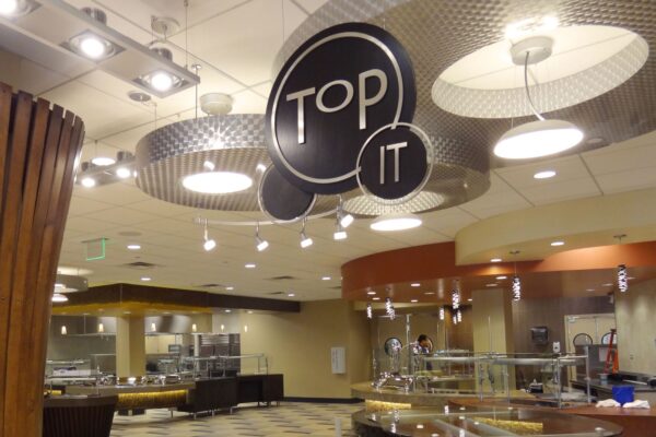 A view of an indoor dining area with circular light fixtures hanging from the ceiling. The sign "Top It" is prominently displayed in the center. The area features checkered floors and various food stations, but no visible people.
