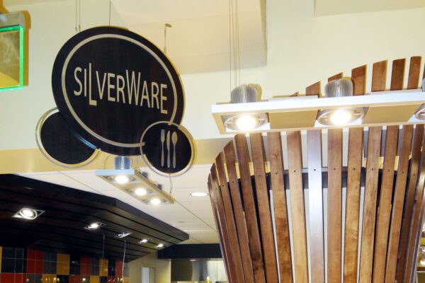 A dining area sign reading "SILVERWARE" hangs from the ceiling, featuring a fork, knife, and spoon icon. In the background are colorful tiles and a ceiling fixture designed with vertical wooden slats.