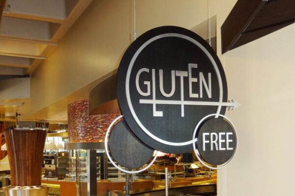 A black circular sign hangs in a restaurant, displaying the words "GLUTEN FREE" in white, modern font. The background of the restaurant shows blurred images of the counters and food displays.